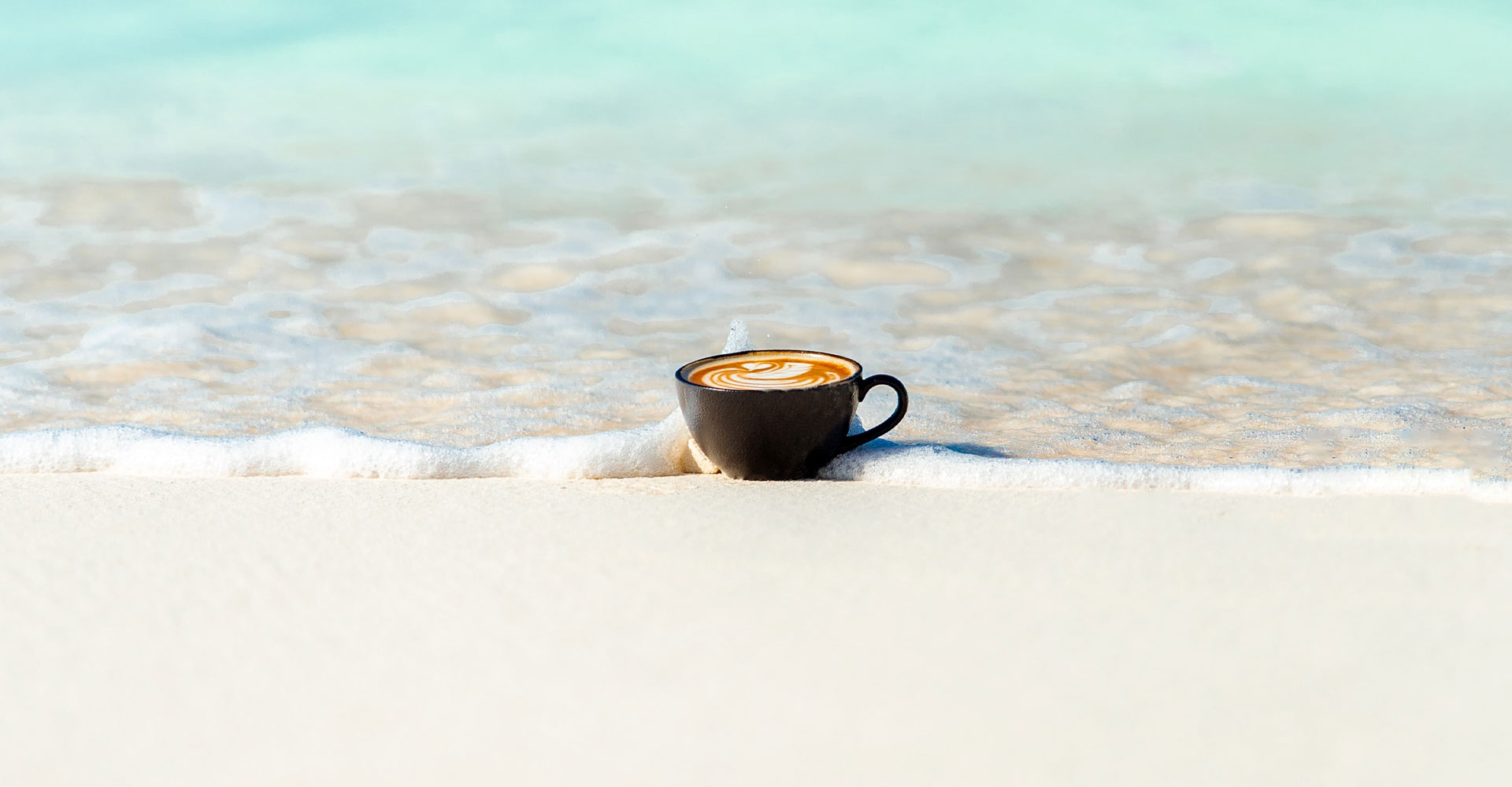 Cappuccino mit Latte-Art-Schwan in schwarze Keramiktasse am Strand mit Meer im Hintergrund