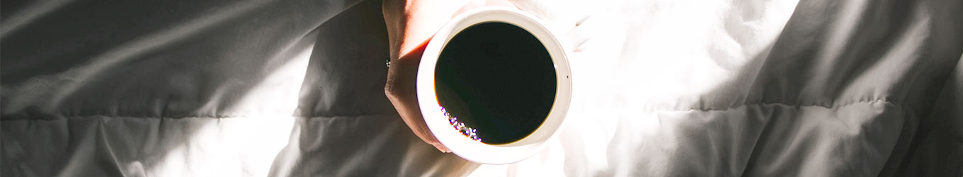 schwarzer Kaffee in weißer Tasse von einer Hand gehalten auf Bettdecke