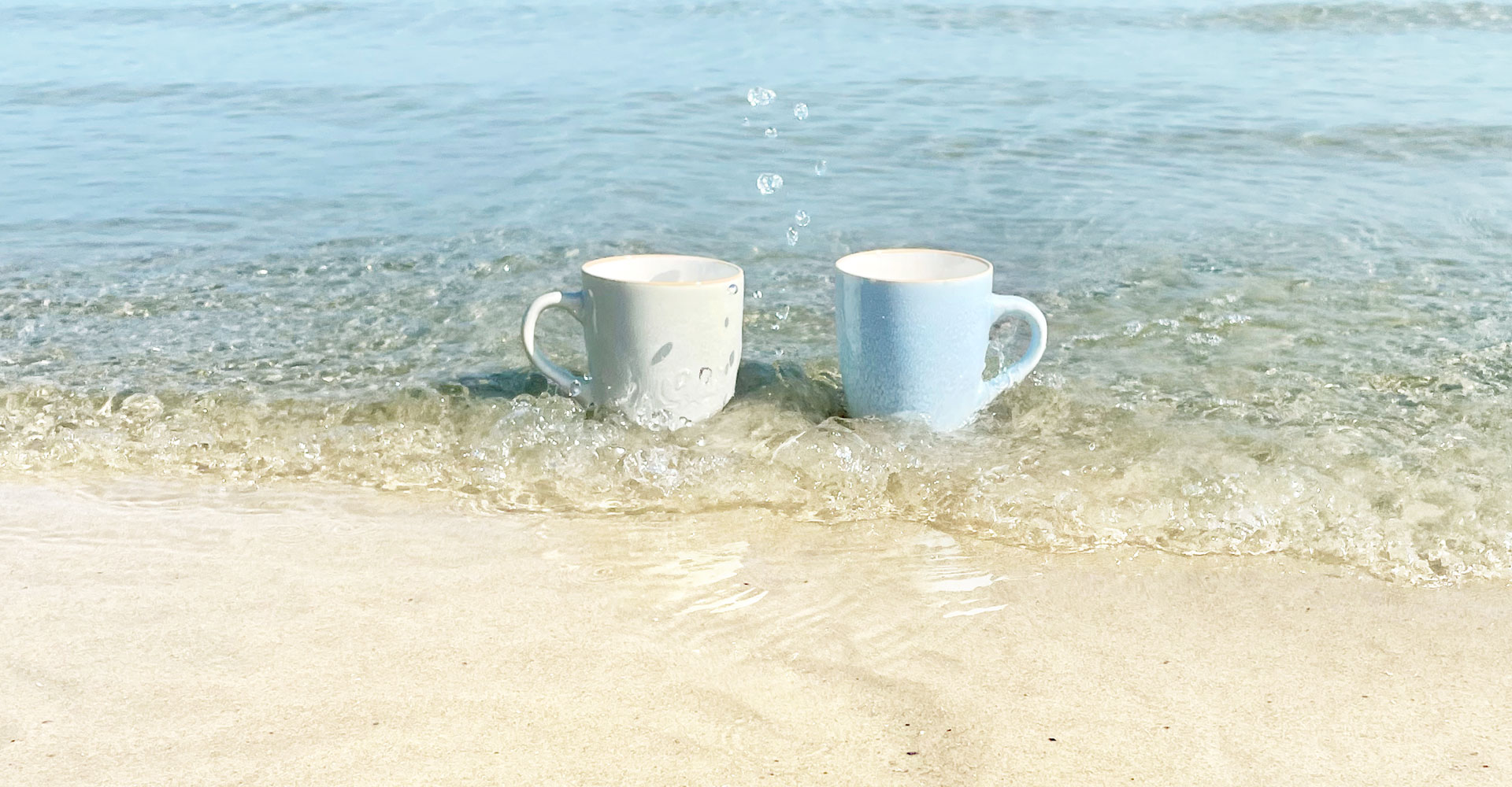 zwei Kaffeetassen stehen am Strand, im Hintergrund die Ostsee